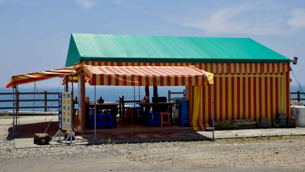 A striped tent eatery offers tables by the coast with Korean signage at the entrance.