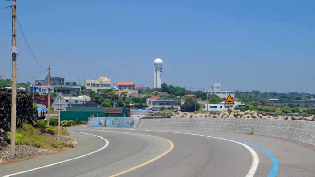 A seawall road with a bike lane curves past a village while a white radar tower stands inland.