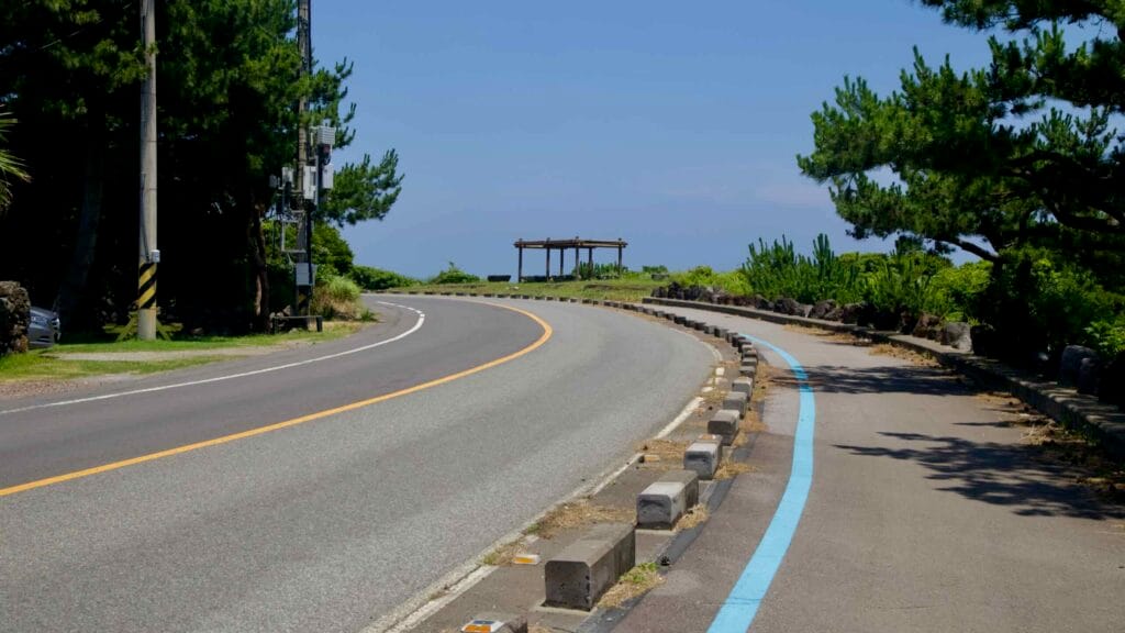 A blue‑striped bike lane curves toward a seaside pavilion on the approach to Seopjikoji.