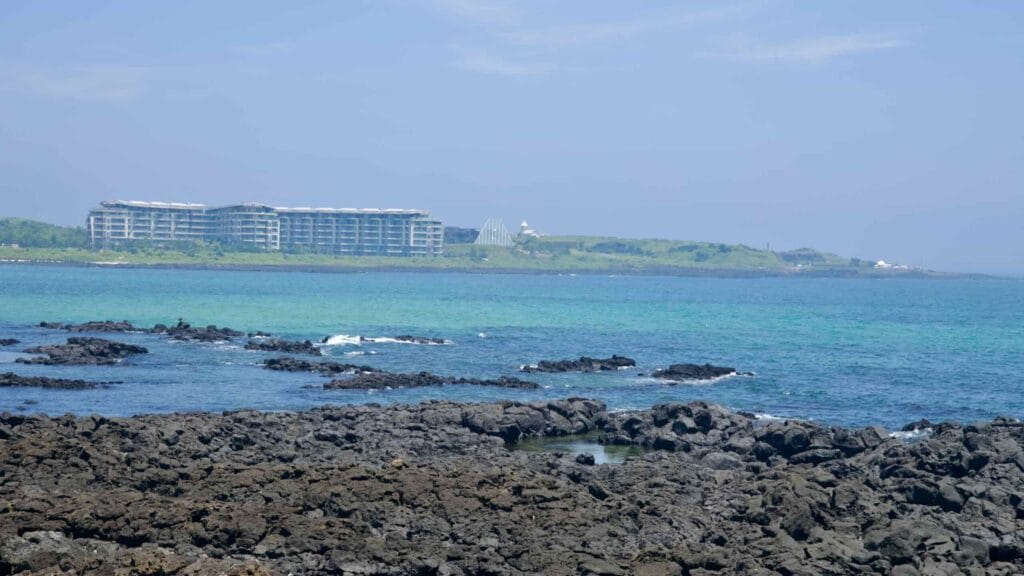 Lava shelves edge turquoise shallows with a resort and glass pyramid on the Seopjikoji shore.