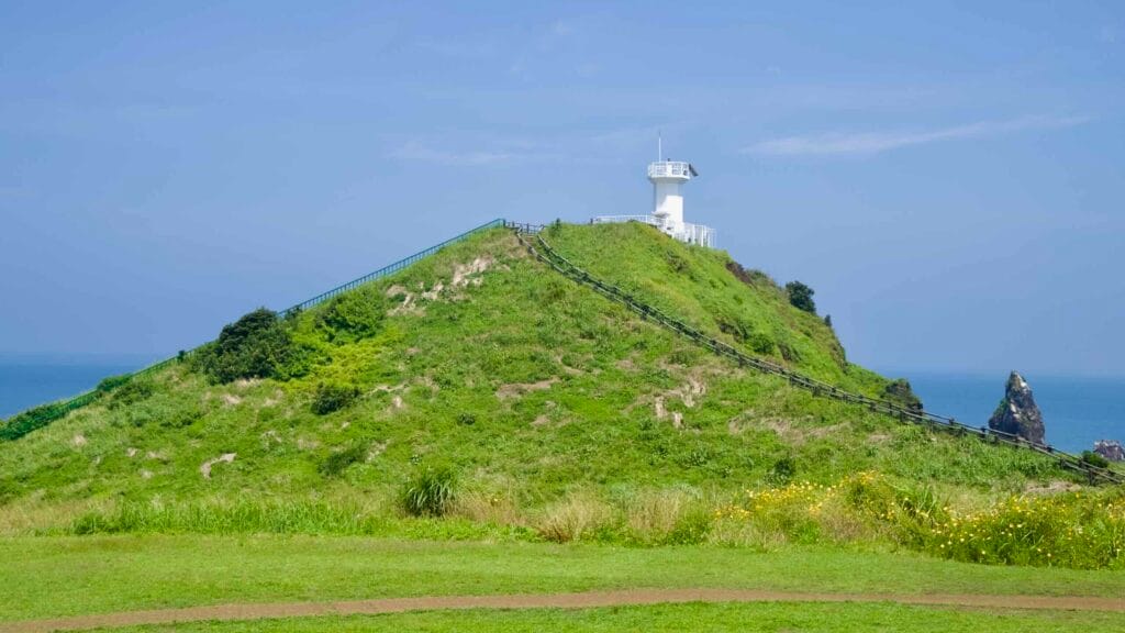Wooden stairs climb to a white lighthouse on the grassy Seopjikoji hill above the cliffs.
