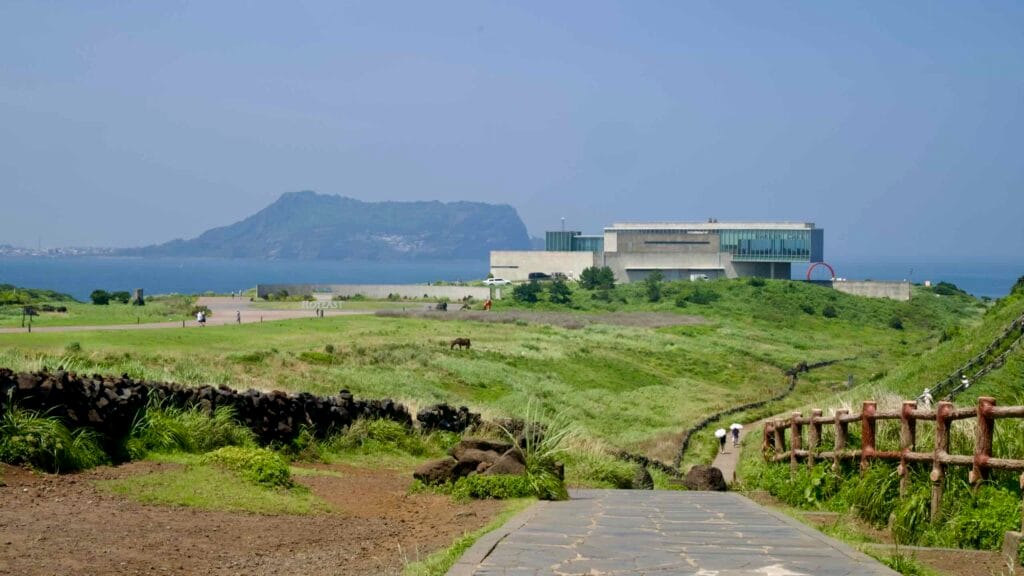 A stone path leads toward the Glass House as horses graze and Seongsan Ilchulbong rises across the bay.