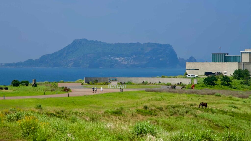 Visitors cross the Glass House lawn with Seongsan Ilchulbong towering across the water.