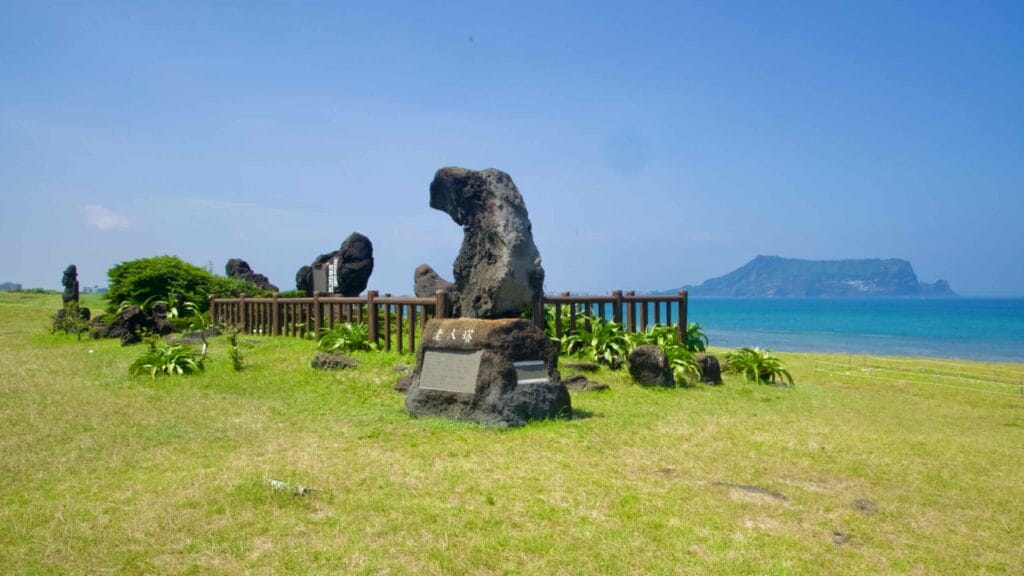 Basalt monuments with plaques stand by the sea at Seopjikoji with Seongsan Ilchulbong beyond.