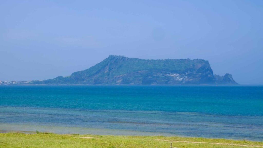 Seongsan Ilchulbong rises across calm water beyond grassy shallows near Seopjikoji.