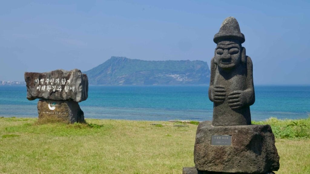 A dol hareubang stone guardian stands near the sea with Seongsan in the distance.