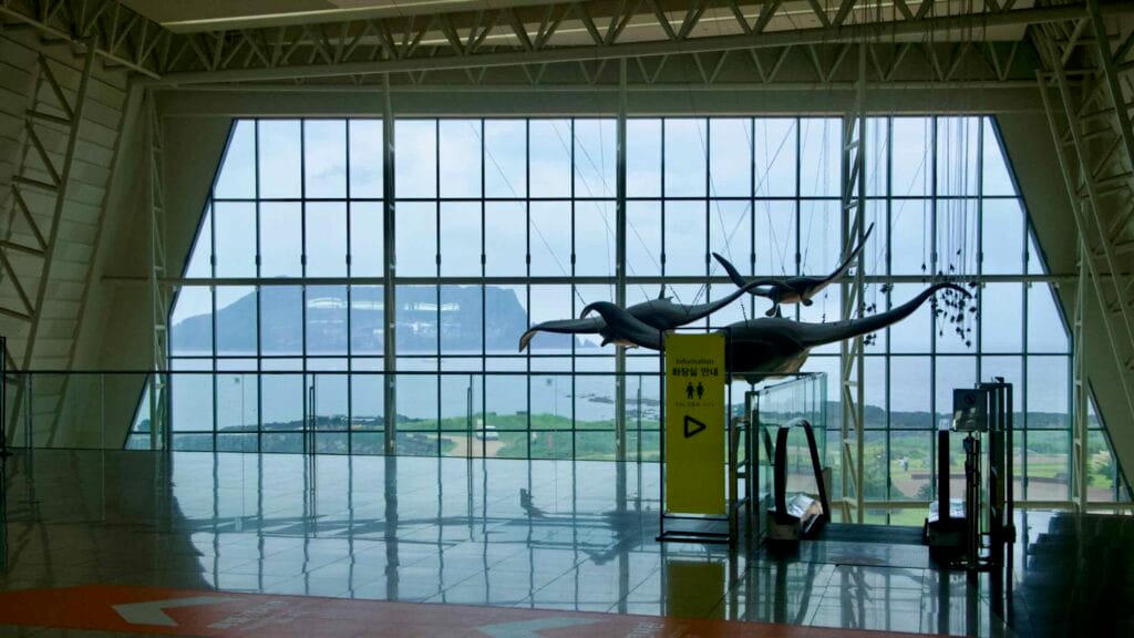 A huge window at Aqua Planet Jeju frames the coast and Seongsan Ilchulbong.