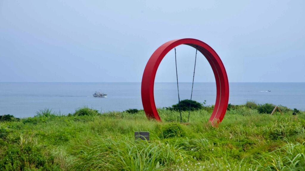 A large red ring sculpture stands in coastal grass at Sopjikoji while fishing boats trace the horizon beyond.