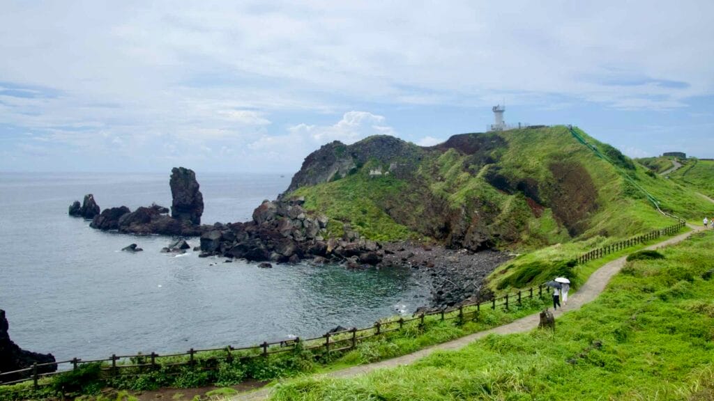 Trails climb the grassy ridge to Sopjikoji Lighthouse as waves meet dark basalt boulders below.