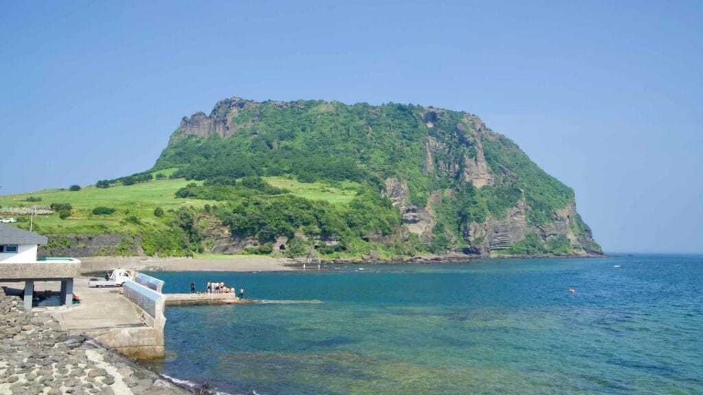 From the harbor edge, Seongsan Ilchulbong towers above green fields and layered cliffs as visitors stand by the water.