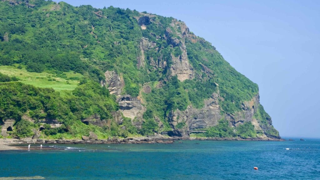 A wide view frames Seongsan Ilchulbong above a cove and turquoise shallows, its grassy apron rolling toward the sea.