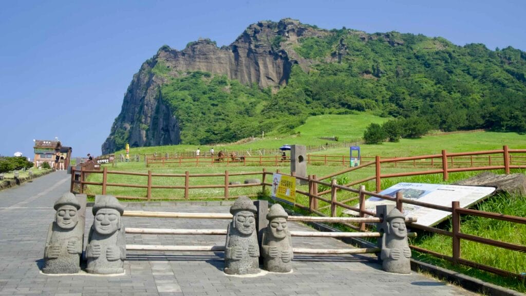 Dol hareubang statues stand at the Seongsan Ilchulbong entrance beside green meadows and walking paths.