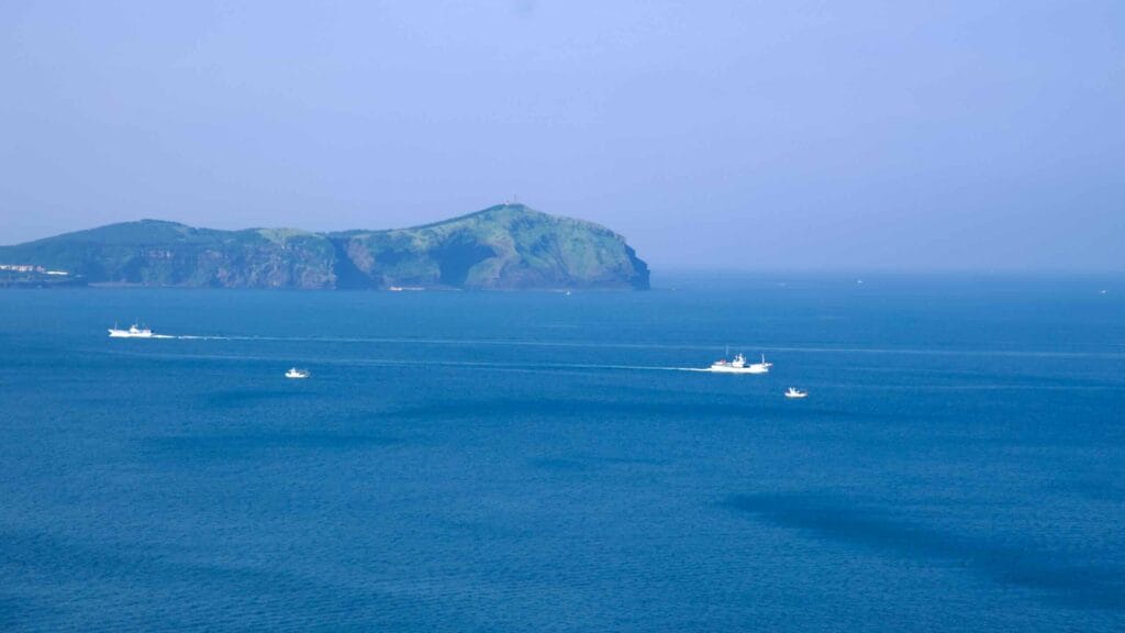 Fishing boats and ferries leave white wakes across the channel toward Udo Island, seen beyond the deep blue sea.