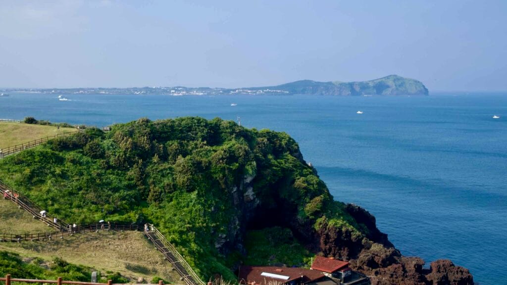 From Seongsan’s trail, a green headland frames a distant view of Udo Island across the calm blue bay.