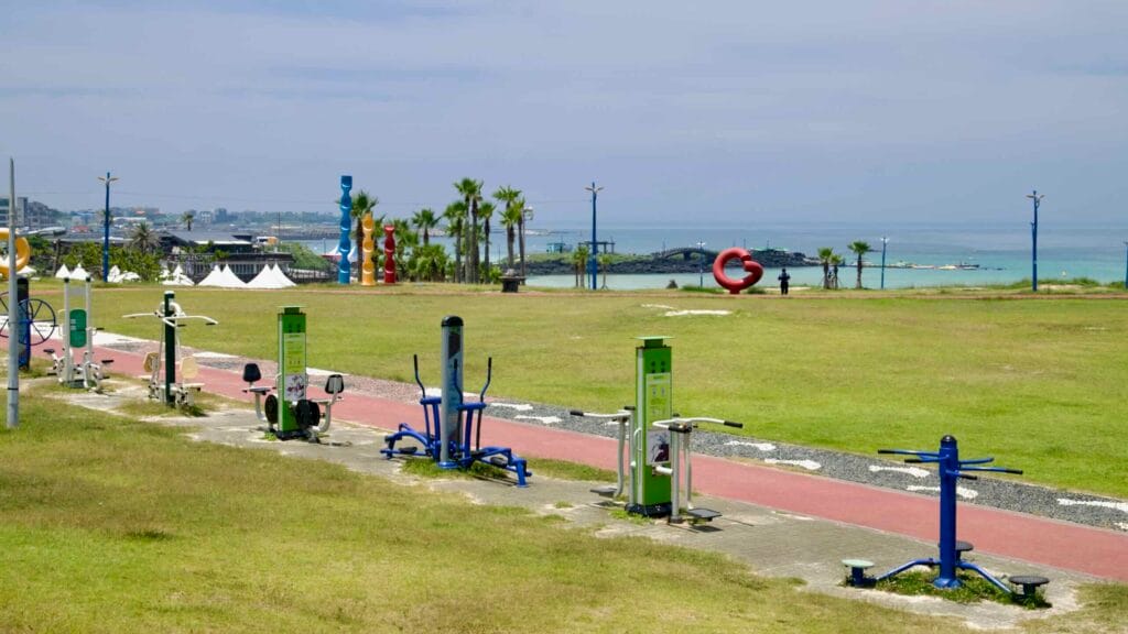 Outdoor fitness machines line a coastal park above Hamdeok Seoubong Beach.
