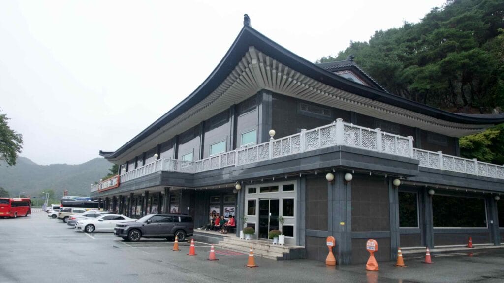 A modern visitor center with sweeping tiled eaves faces a wet parking lot at Mungyeong Saejae, buses and cars lined up in the rain.