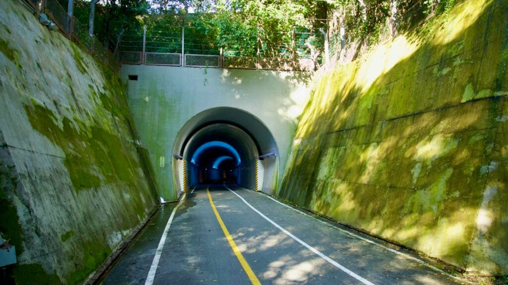 At the opposite portal, blue lighting outlines Saekhyeon Tunnel’s arch as dappled sunlight falls across the path.