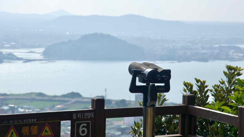 A binocular viewer stands at Viewpoint 6 on Seongsan Ilchulbong, aimed toward the lagoon.