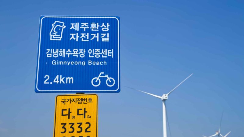 A Gimnyeong cycling sign stands with turbines beyond a clear sky.