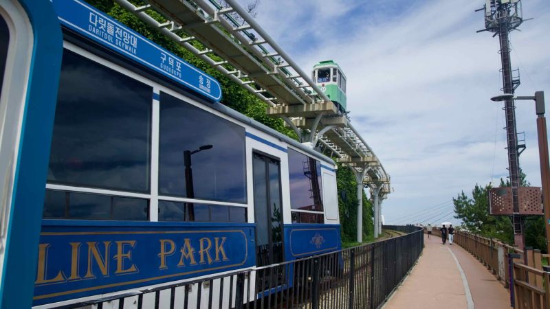 Beach train by the boardwalk as a green sky‑capsule passes overhead.