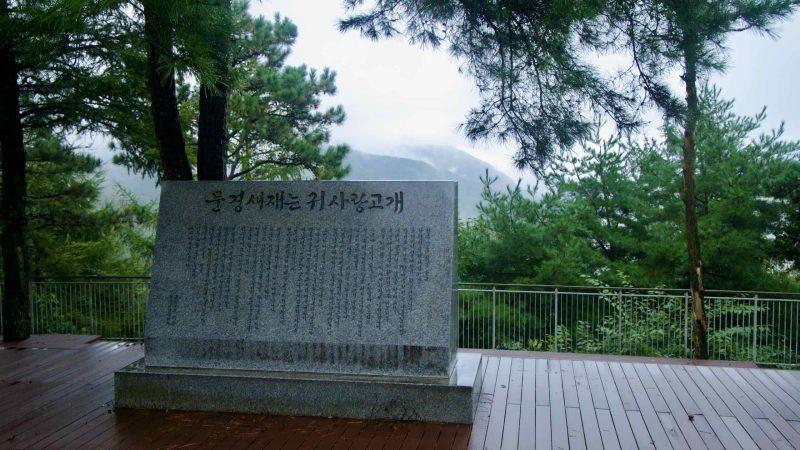 A granite stele recounts Mungyeong Saejae history on a rainy wooden deck.