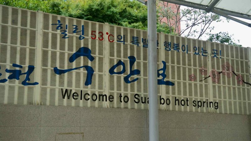 An entrance wall reads “Welcome to Suanbo hot spring” beside calligraphy and blossoms.