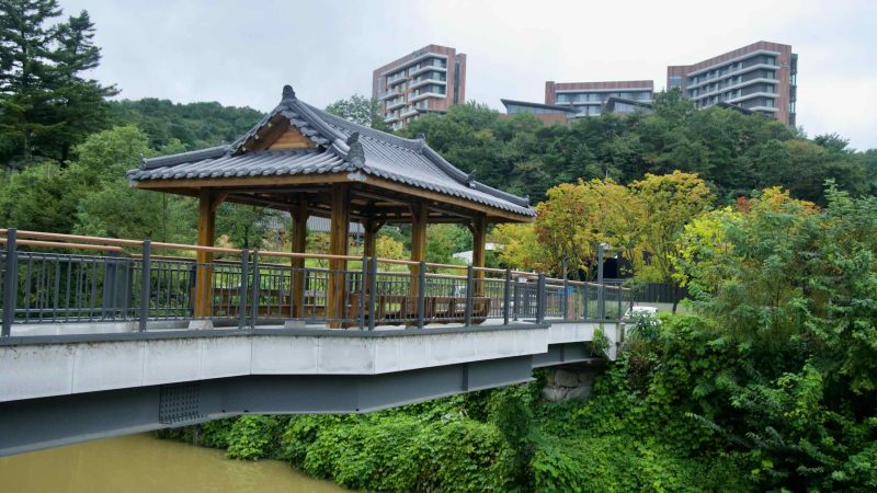 A tile-roofed wooden pavilion sits on a bridge with resorts beyond the trees.