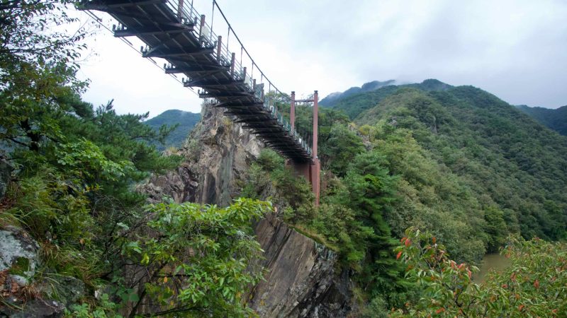 From the ridge, the bridge crosses between cliffs above the Dalcheon with misty mountains beyond.