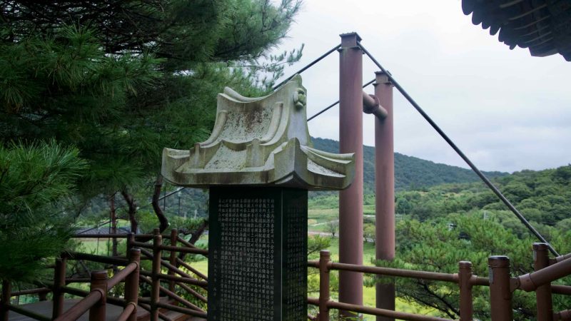 A roof-topped stone stele stands near the bridge towers and cables above green hills.