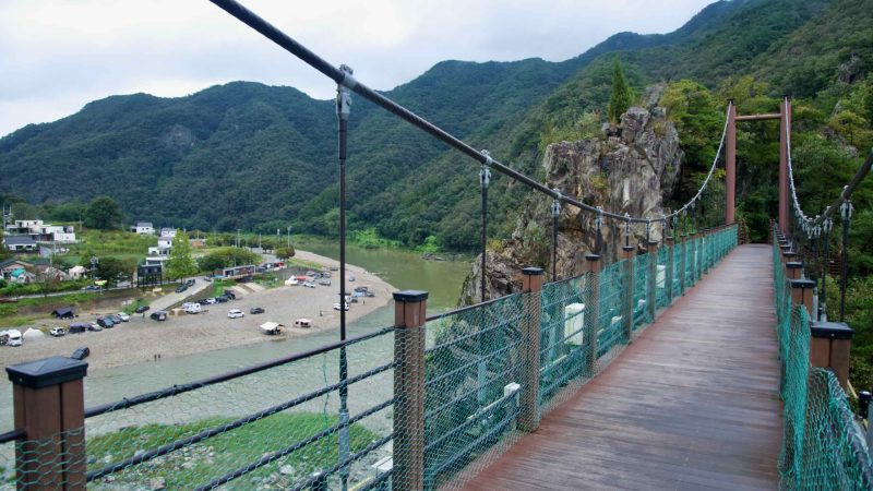Standing on the Cloud Bridge, visitors look toward a gravel-bar campground at the Dalcheon’s bend, where cars and tents cluster.