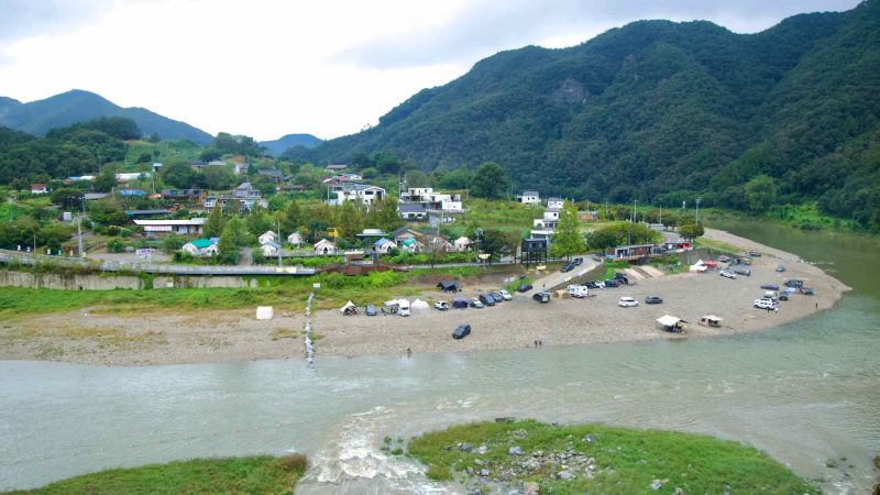 Across the Dalcheon, a riverside village and rows of tents and vehicles line the gravel bar near Sujupal Peaks beneath cliffs.
