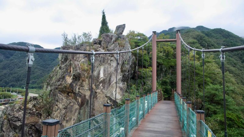 A bridge tower stands on the cliff edge beside a narrow crag topped with a lone tree, where cables carry the deck to the ridge.