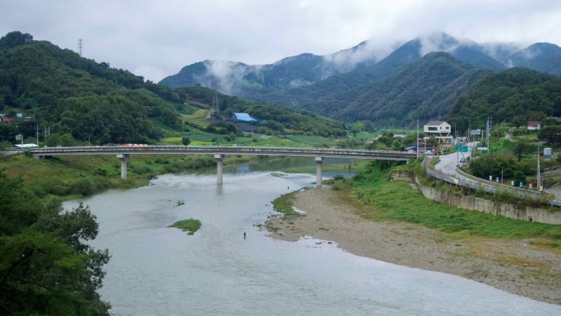 A concrete highway bridge crosses the Dalcheon upstream, with fog clinging to ridges and scattered farms and houses.