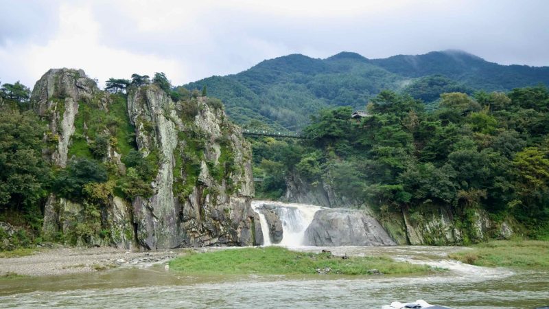 A twin-cascade pours over bedrock into the Dalcheon while the Cloud Bridge and a hilltop pavilion peek through pines along the rim.