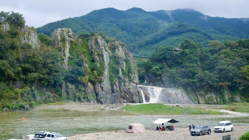 Cars and tents line the riverside gravel bar facing Kal Rock Falls, with the suspension bridge threading between the cliffs above.