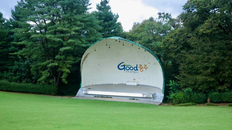 An open-air bandshell with a curved roof stands on the lawn at Tangeumdae Park, small stage lights hanging beneath.