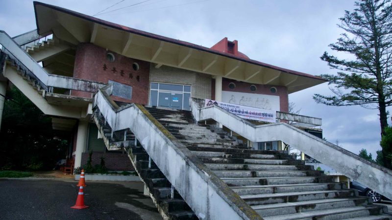 Broad concrete stairs rise to the cultural hall at {loc}, their damp steps leading past a banner and brick wall.