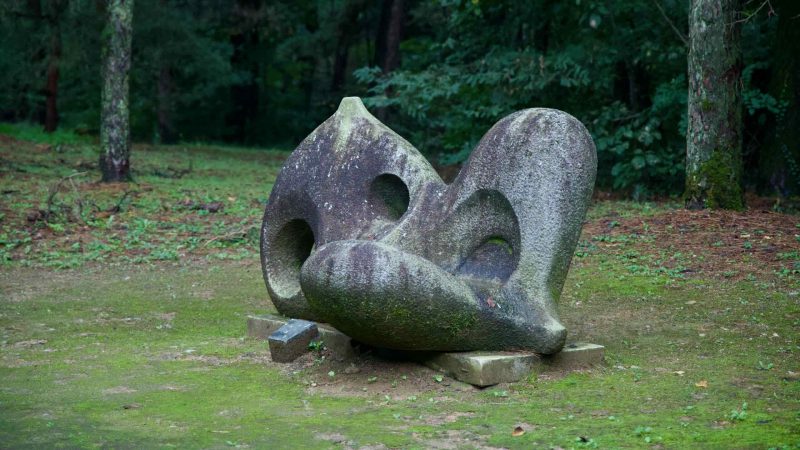 An abstract stone sculpture rests on mossy ground at {loc}, its rounded forms and voids catching the soft light.