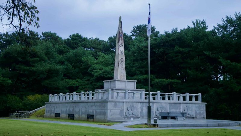 A granite memorial tower with Korean inscription rises beside a flagpole at Tangeumdae Park, honoring General Sin Rip.