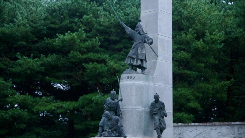 Bronze figures of patriots stand at a granite obelisk in Tangeumdae Park, one raising a flag beside carved reliefs.