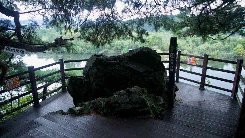 A dark boulder anchors a compact overlook deck at Tangeumdae Park above the river, with caution signs on the rail.