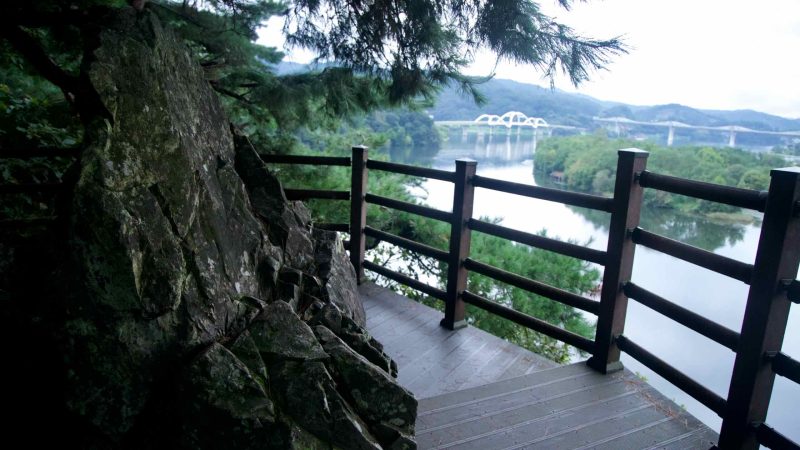 A rocky ledge and railing frame a clear view from Tangeumdae Park across the South Han River and wooded banks.