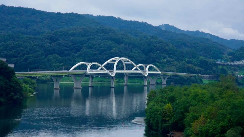 Curving white arches of a road bridge span the South Han River near Tangeumdae Park, framed by distant wooded hills.