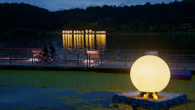 A glowing moon sculpture with rabbits floats at Tangeum Lake as cyclists pass below.