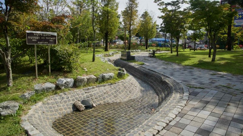 A stone-lined water channel winds through a small urban park in downtown Chuncheon.