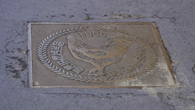 A square bronze pavement plaque labeled Dakgalbi Street with a rooster emblem and 1968 is set into the lane.