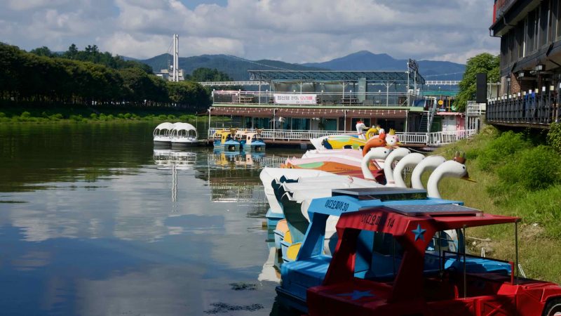 Colorful swan and animal-shaped pedal boats line a dock on Gongji Stream near cafes and low hills, with calm water reflecting them.