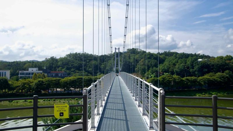 Pedestrians cross Chuncheon’s Sairo 248 suspension bridge above Uiam Lake and low forested hills.