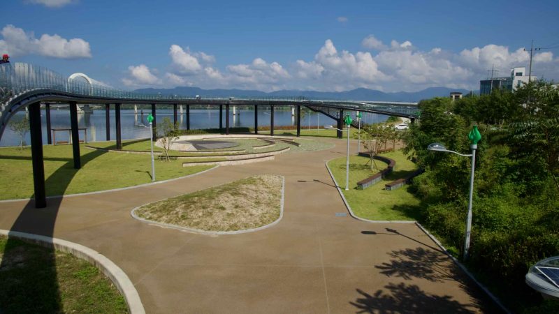 Paths, lawns, and elevated glass walkways spread across Munhwa Gwangjang Forest beside Lake Uiam.