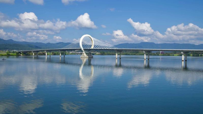 Cyclists and walkers share a lakeside park path in Chuncheon beside open lawns and water.